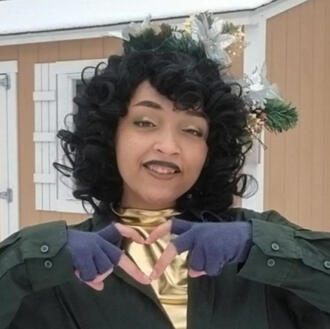 A close up of a person with feminine features, cosplaying as Lup Taaco. They are tan, with curly black hair, and are dressed in winter clothes, making a heart with their fingers.